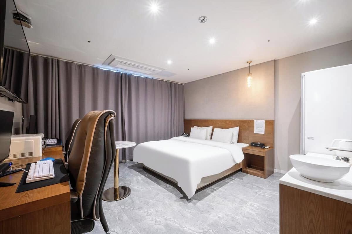 Habitación de hotel con cama, escritorio con computadora, silla, mesita de noche con lámpara y baño con lavamanos y espejo.