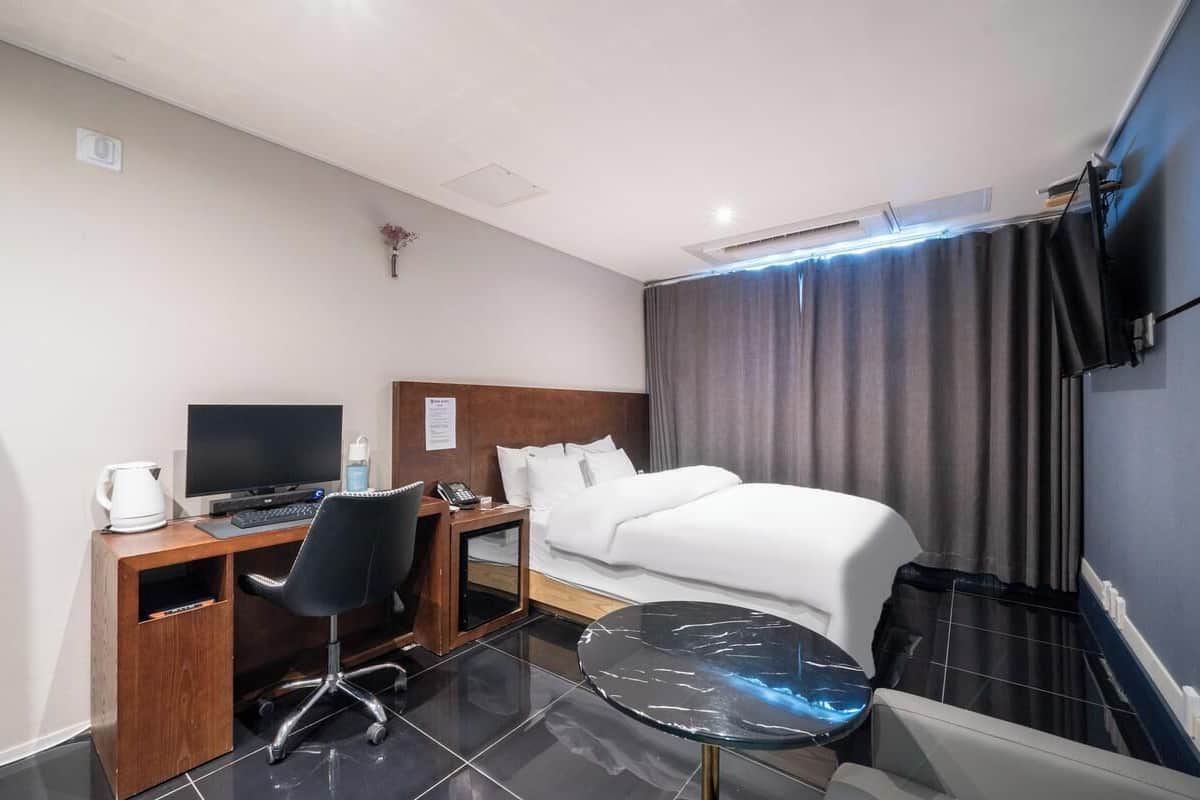 Una habitación de hotel con una cama, un escritorio con computadora, una silla, una mesita y un televisor montado en la pared.