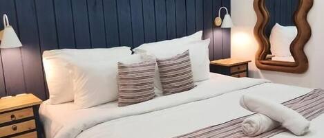 A neatly made bed with white bedding and striped pillows, a wooden nightstand with a lamp, and two framed photographs on the wall.