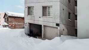 Ein mehrgeschossiges Betongebäude mit Garagentor, einem Holzbau im Hintergrund und blauem Himmel.