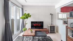 Ein modernes Wohnzimmer mit Flachbildfernseher, der 'NETFLIX' anzeigt, einem Essbereich mit Tisch und Stühlen, einer großen Topfpflanze und einer Küchenzeile mit Kühlschrank und Mikrowelle.