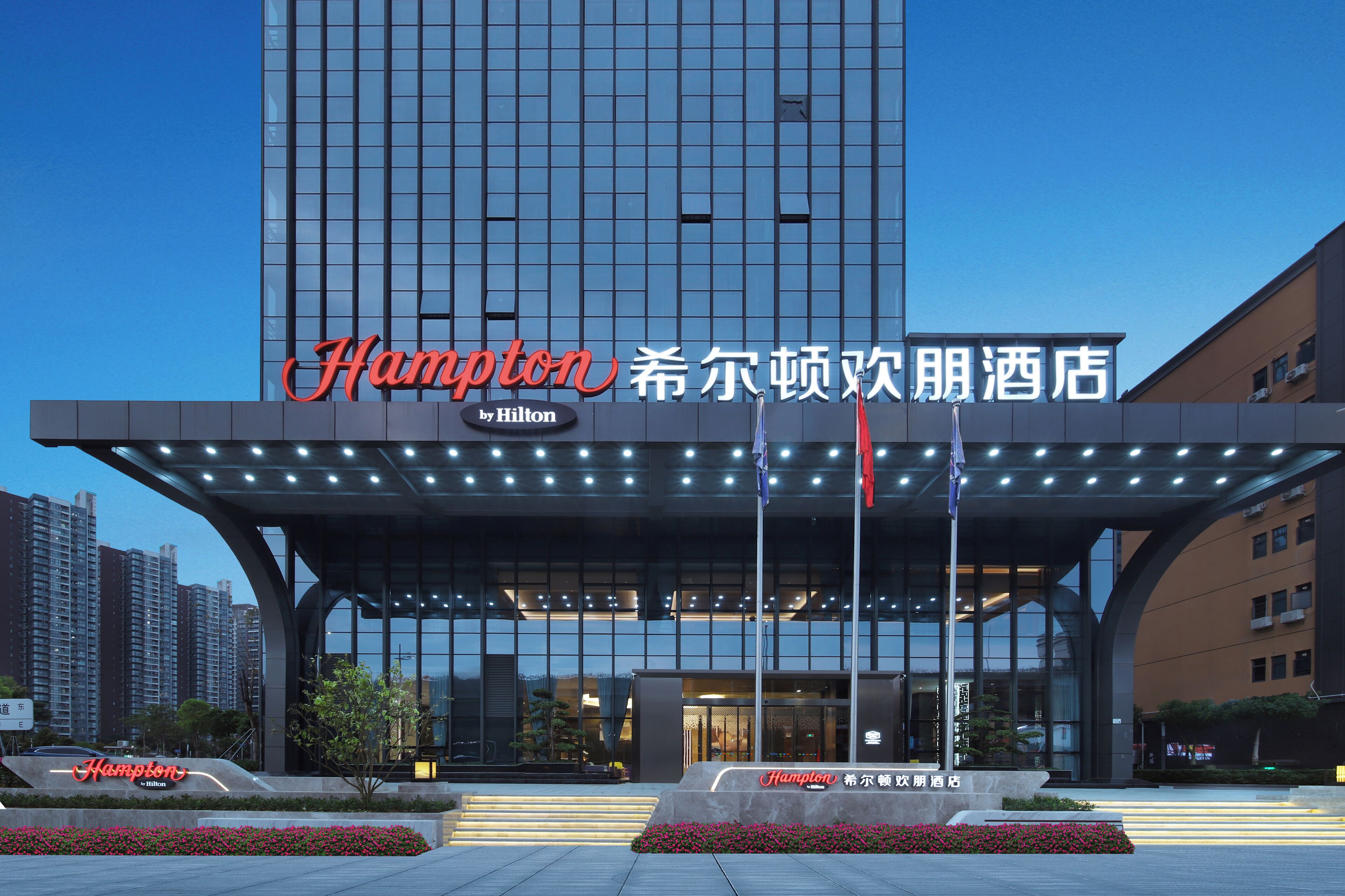 Um hotel moderno com uma placa chamativa da 'Hampton by Hilton', amplas janelas de vidro e uma entrada bem iluminada.