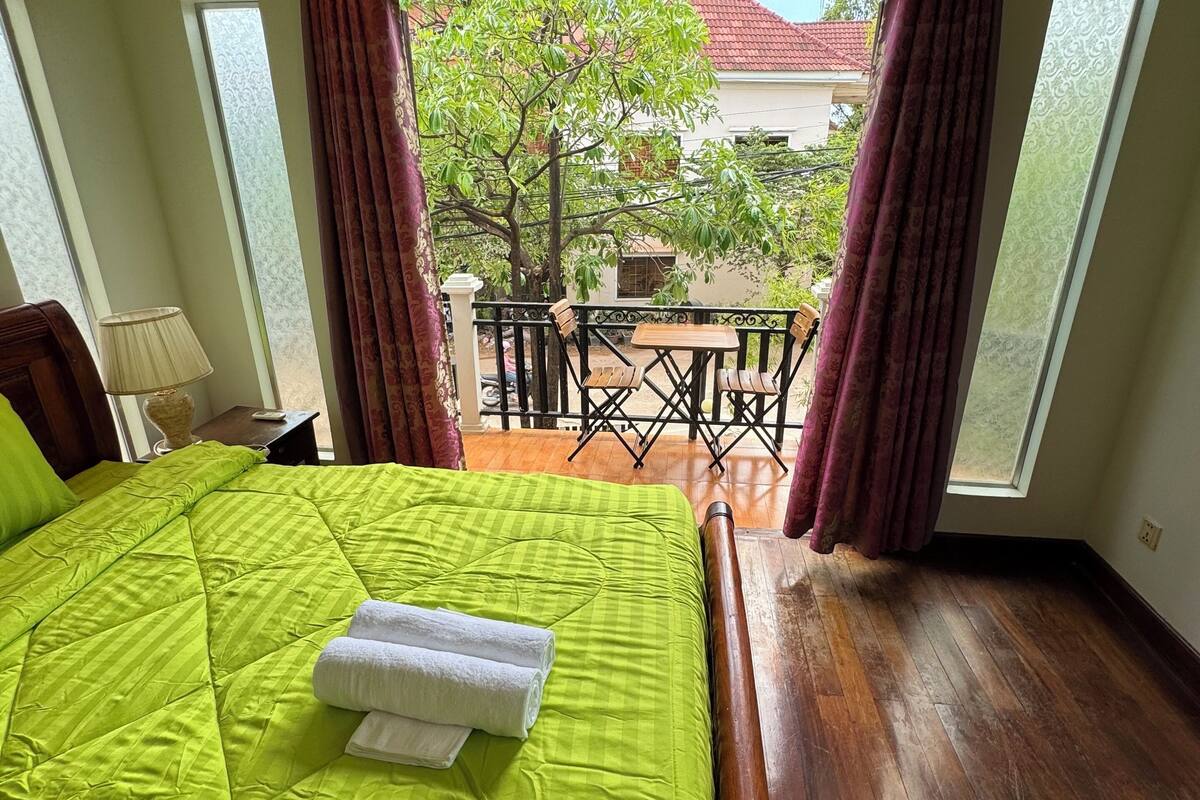 A hotel room with a large bed, a balcony with a table and chairs, and a view of the outside.