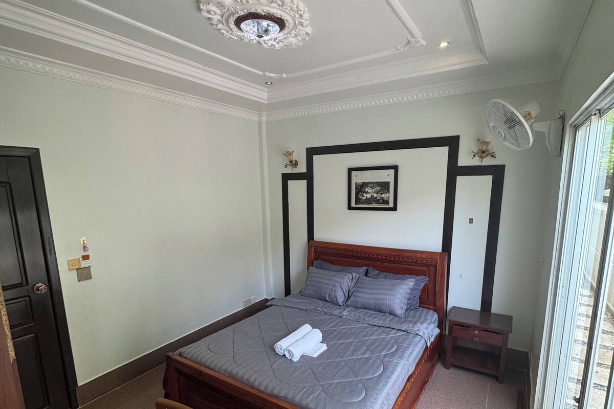 A bedroom with a bed, a chair, a small table, a fan, and a picture on the wall.