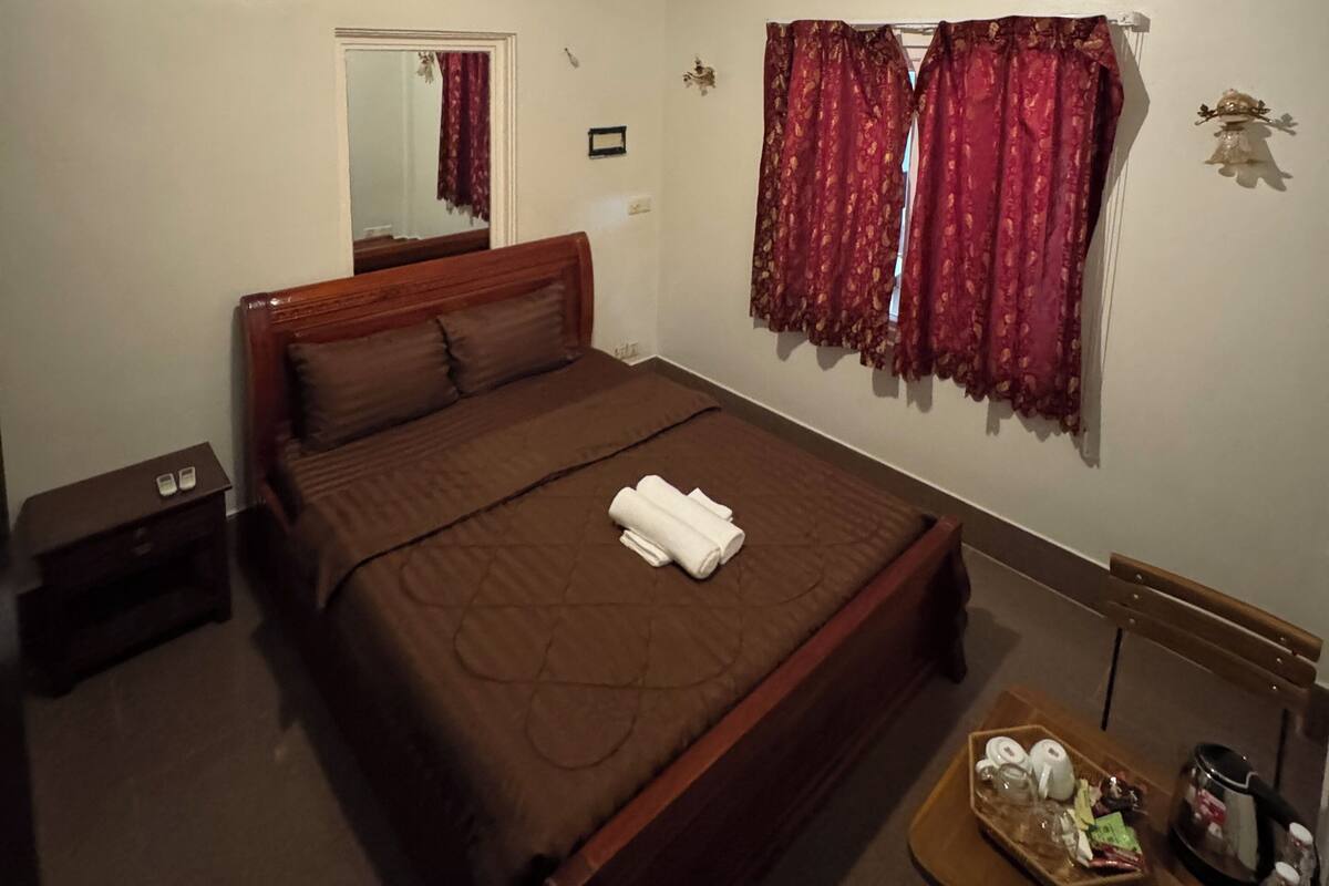 A bedroom with a large bed, a wooden headboard, a nightstand, a table with toiletries, and a window with curtains.