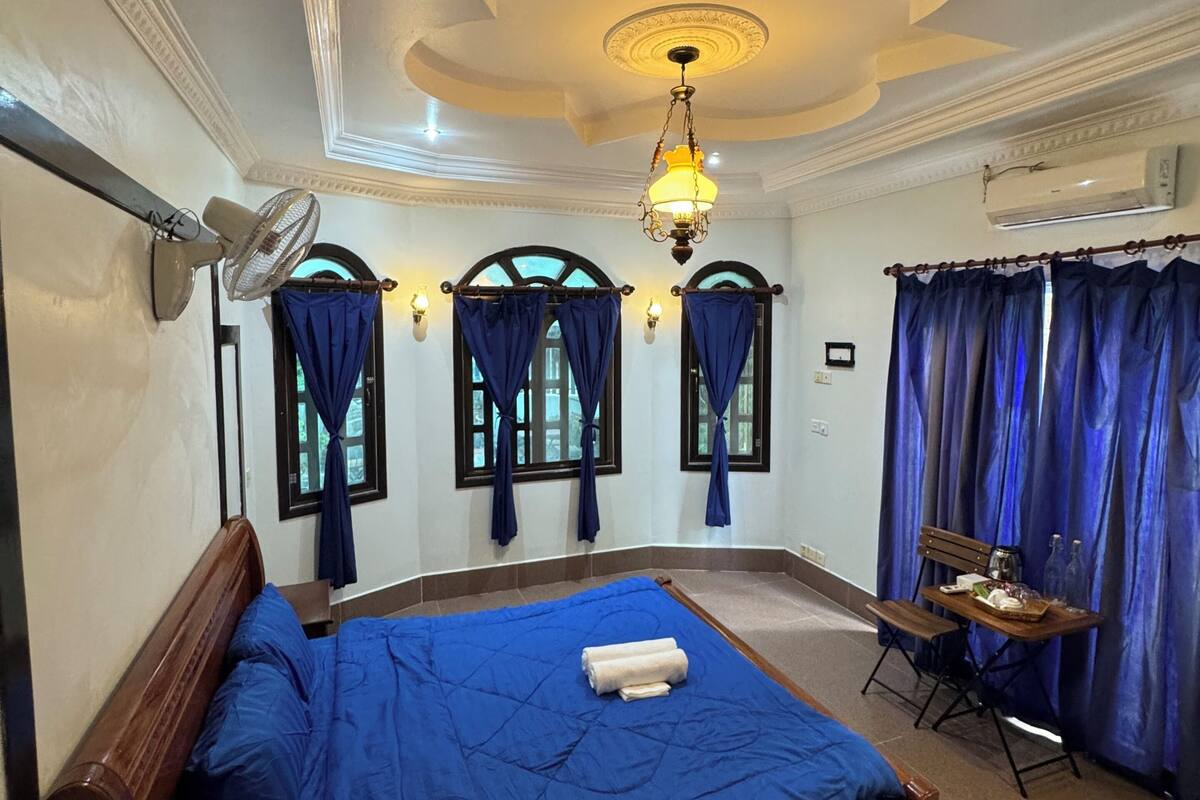 A hotel room with a bed, a desk with a chair, a standing fan, a chandelier, and a window with curtains.