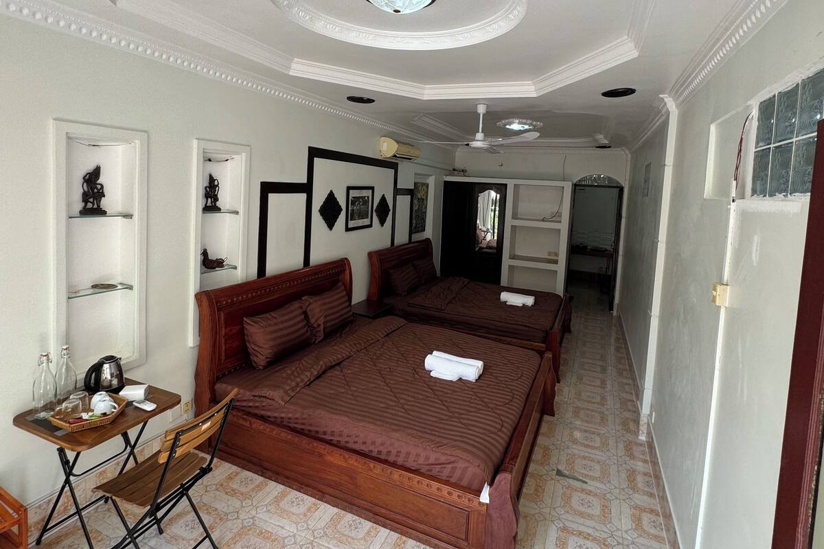 A hotel room with a large bed, a small table with a chair, a ceiling fan, and a tiled floor.