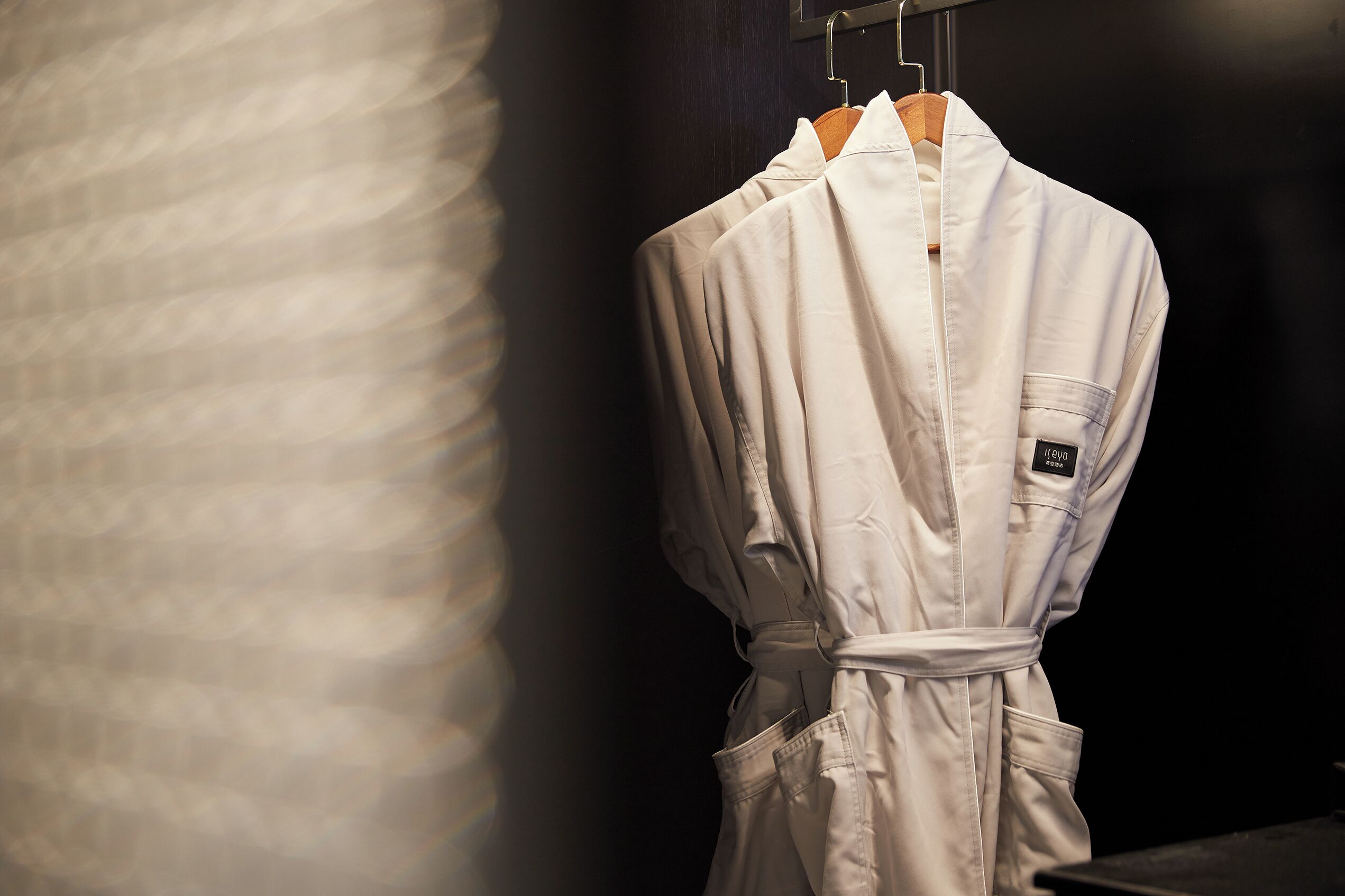 Une robe blanche est suspendue à un cintre en bois dans une pièce faiblement éclairée, dont le mur présente une texture particulière.