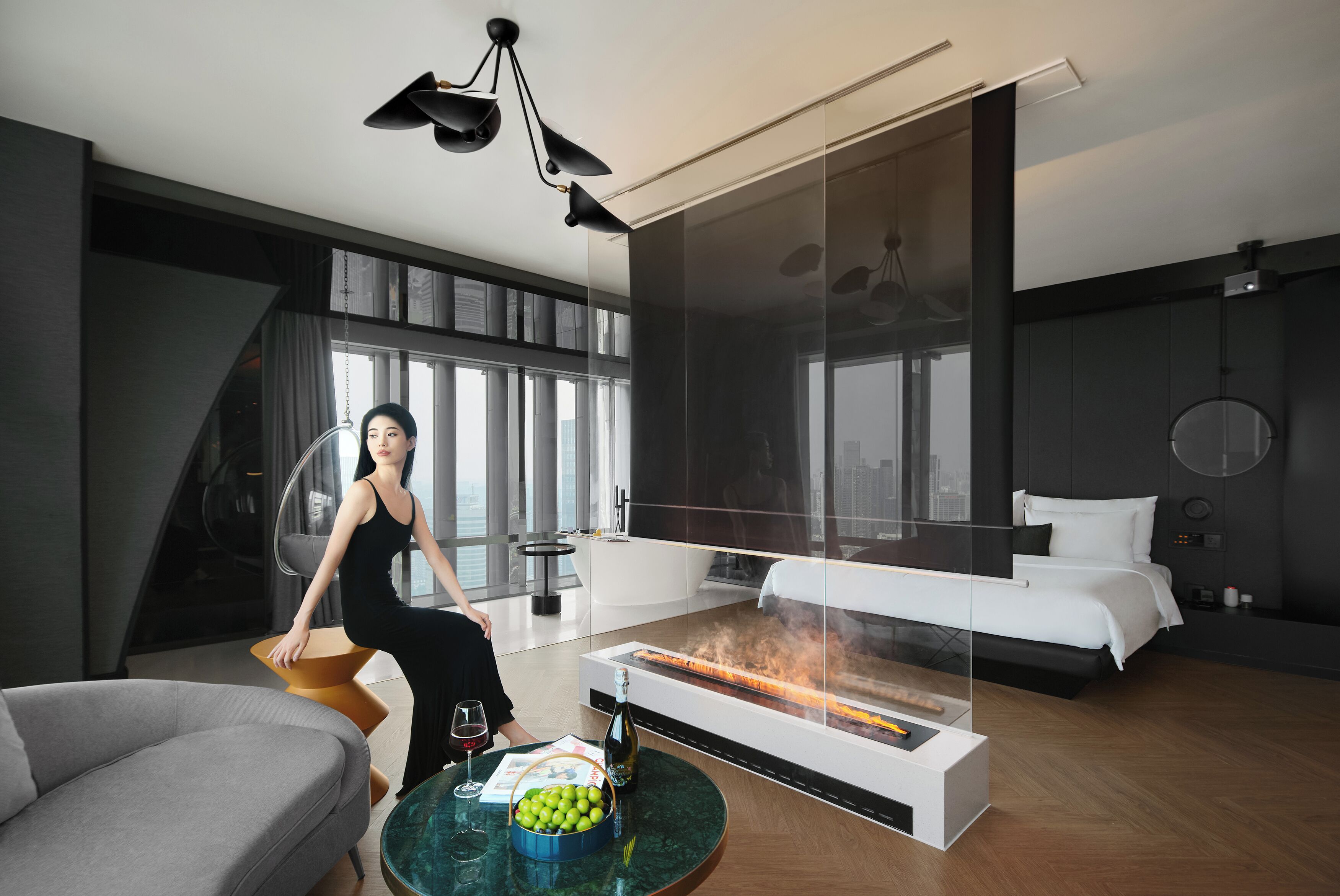 A modern hotel room with a glass fireplace, a sofa, a dining table with a bowl of fruit, and a bed.