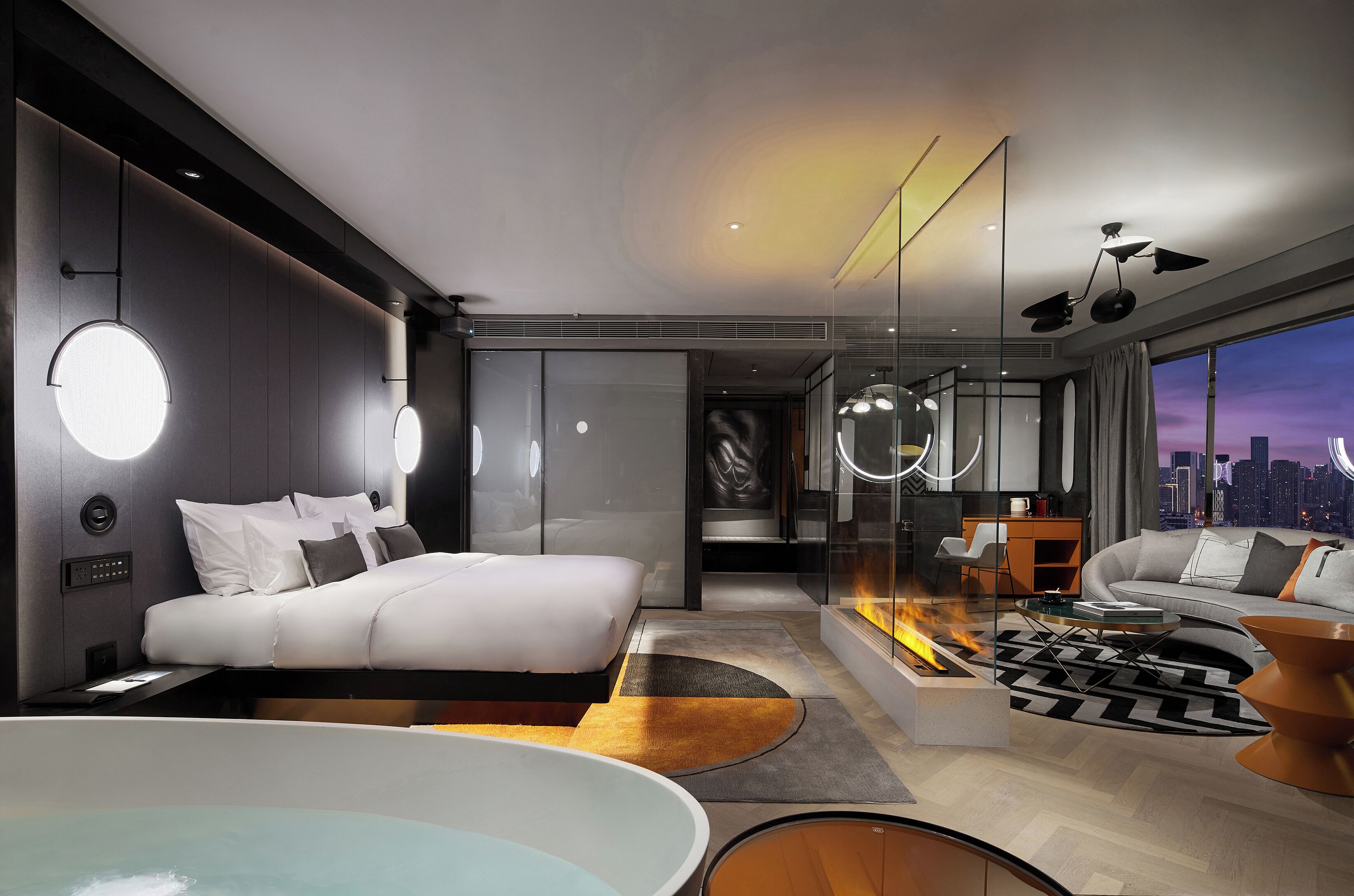 A modern hotel room with a bed, a sitting area, a large window with a city view, a bathtub, and a fireplace.