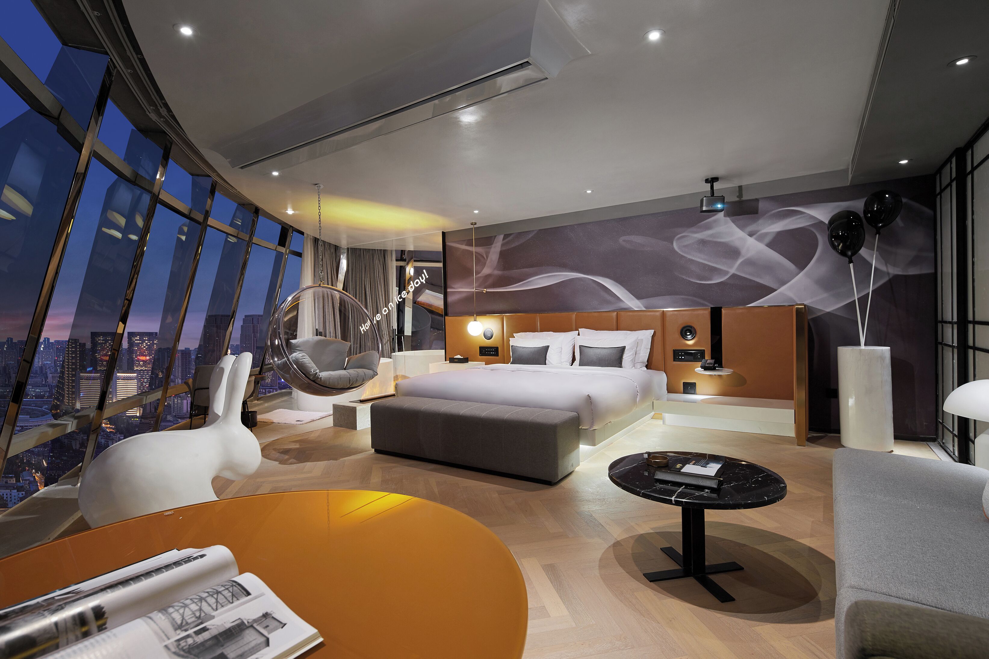 A modern hotel room with a large bed, a sofa, a dining table, and a view of the cityscape.