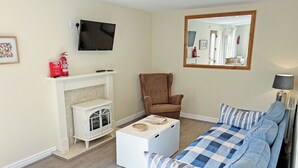 A living room with a white fireplace, a brown armchair, a blue sofa, and a flat-screen TV mounted on the wall. - Elder Cottage - UK51371 (Caldwell, Richmond)