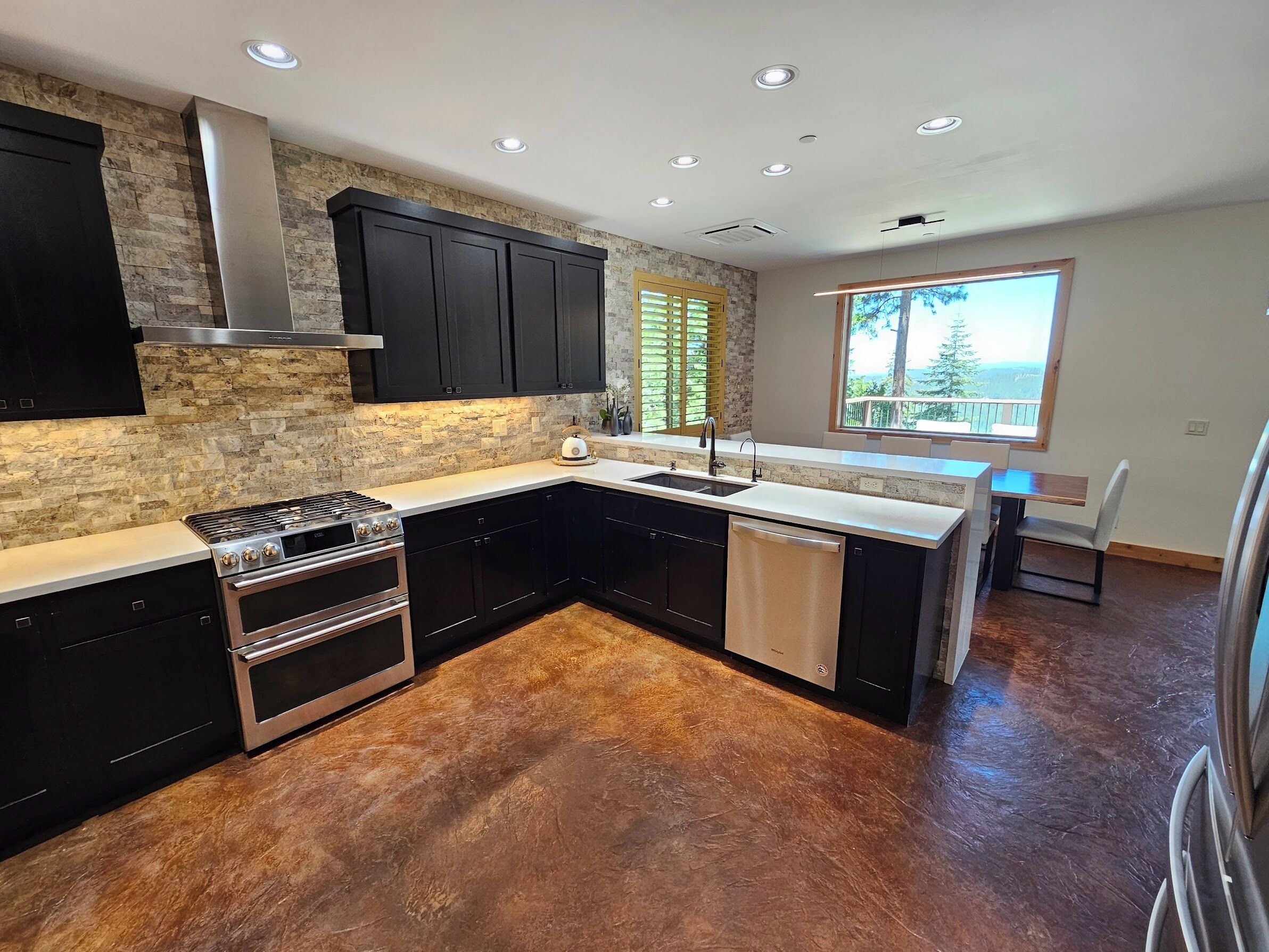 A modern kitchen with a stone backsplash, black cabinets, stainless steel appliances, and a large window offering a view.