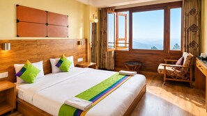 A hotel room with a large bed, wooden headboard, a view through a window, a chair, and a nightstand. - Dekeling Hotel (Darjeeling)