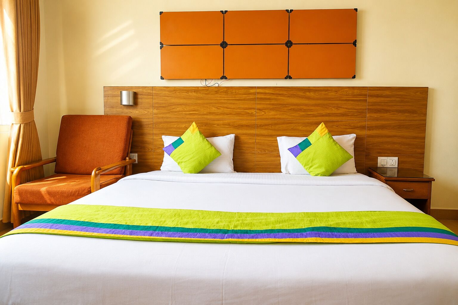 Habitación de hotel con cabecera de madera, cama con ropa blanca y almohadas coloridas, mesita de noche de madera y una silla.