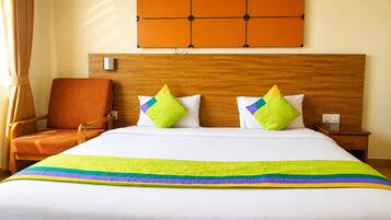 Habitación de hotel con cabecera de madera, cama con ropa blanca y almohadas coloridas, mesita de noche de madera y una silla.