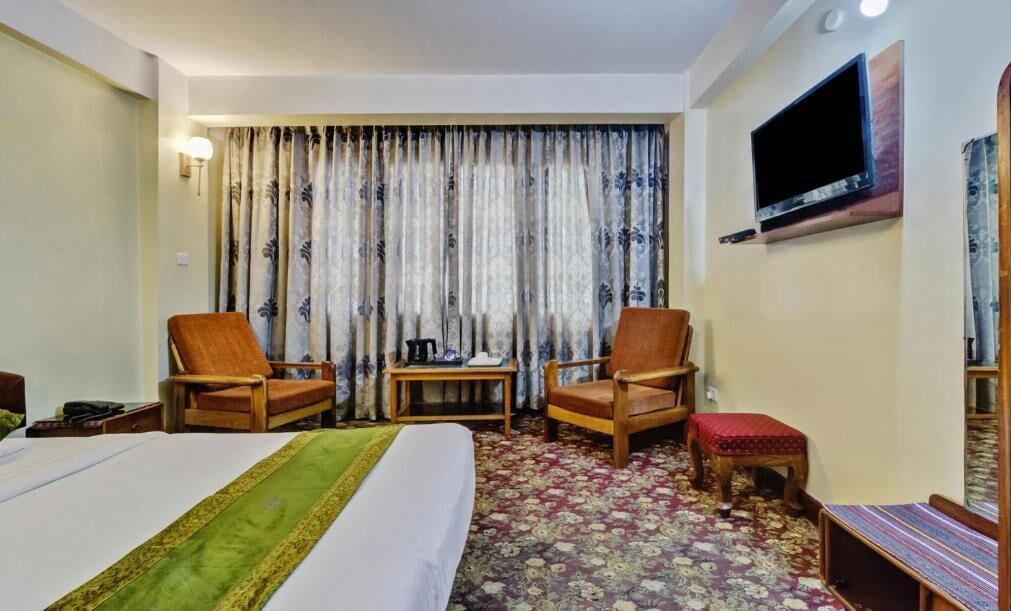 A hotel room with a bed, two wooden chairs, a small table, a TV mounted on the wall, and a patterned carpet.