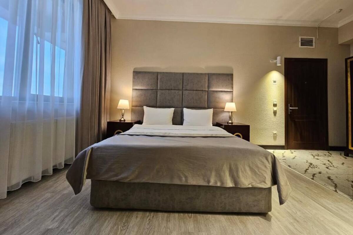 A hotel room with a large bed, two bedside tables with lamps, a headboard, a door, and a window with curtains.