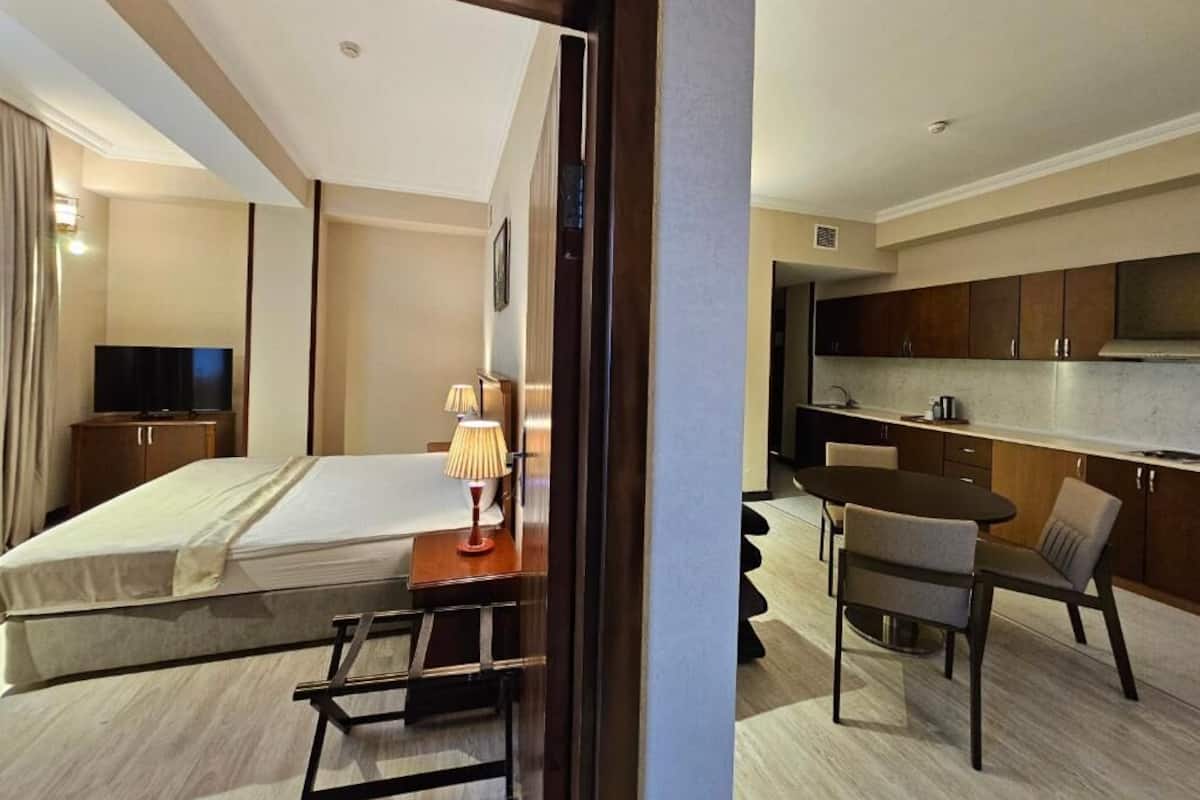 A hotel room with a bed, bedside table, lamp, and a view of the kitchen area.