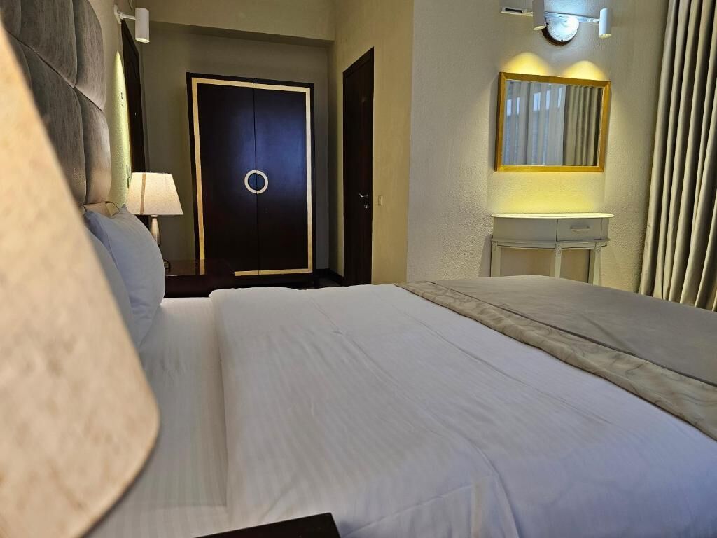 Una habitación de hotel con cama, lámpara de noche, espejo, puerta y una mesita.