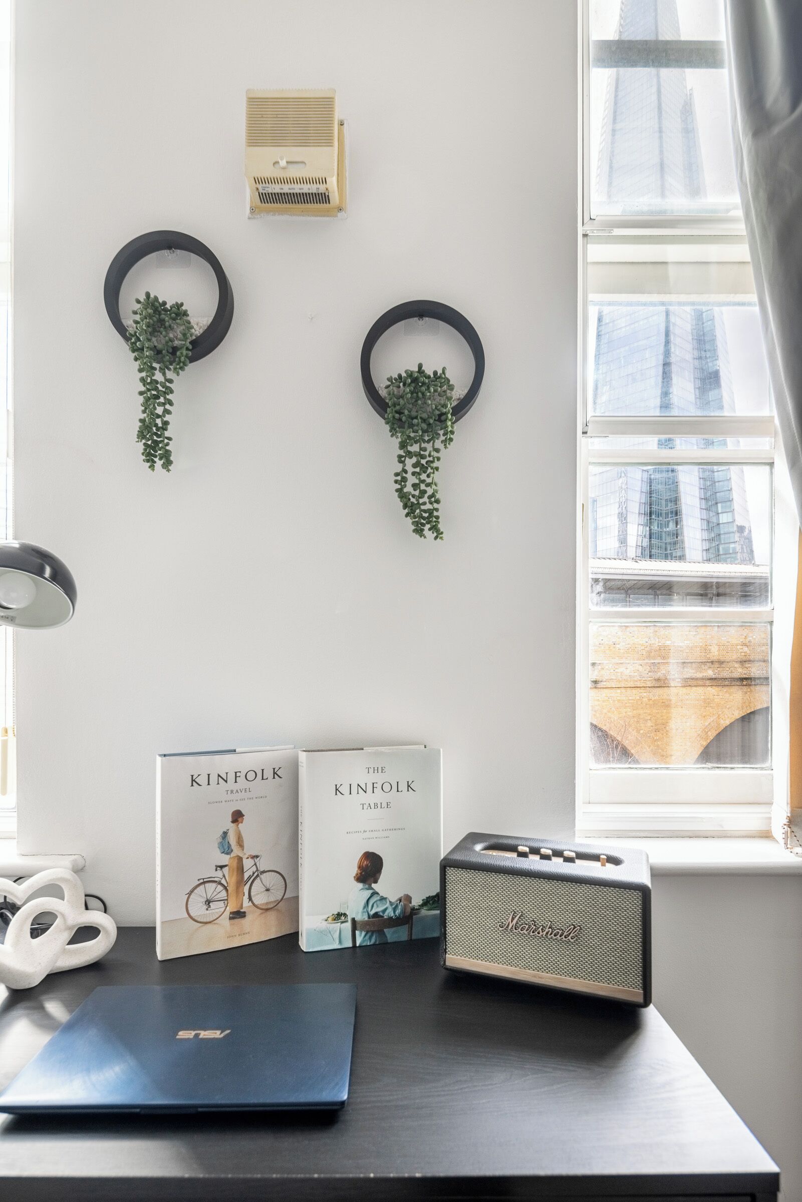 Een bureau met een laptop, een Marshall-speaker en twee boeken met de titel 'KINFOLK' aan een muur met uitzicht op een raam.