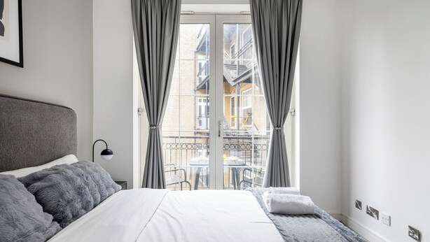 A bedroom with a bed, grey curtains, a window with a view of buildings, and a small table with chairs outside. - Luxury 2 Bedroom Apartment near London Bridge • Sleeps 6 • Tube Across Street (London)