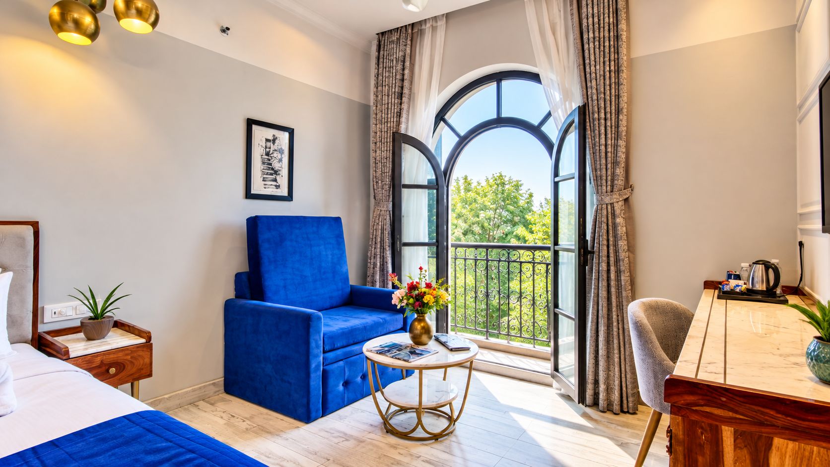 A hotel room with a bed, a blue sofa, a round table with a vase of flowers, and a balcony with a view of greenery.