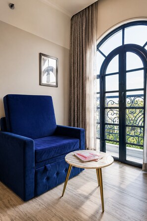 Une pièce avec un canapé bleu, une petite table ronde sur laquelle repose un magazine, une grande fenêtre en arc de cercle offrant une vue dégagée, et un tableau encadré accroché au mur.