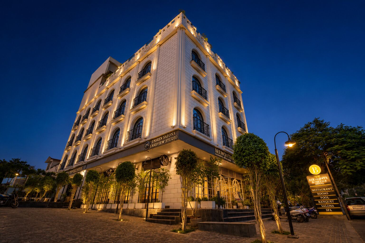 Un hotel de varios pisos con una entrada destacada, señalización que indica 'UNSEEN SALONA LAKEVIEW STUDIO' y 'BELMONTE HOTEL', y una fachada bien iluminada.