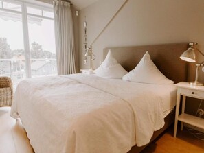 A bedroom with a large bed, two bedside tables with lamps, a window with curtains, and a view of buildings outside. - Heikendorf Penthouse Suite by Belvilla (Heikendorf)
