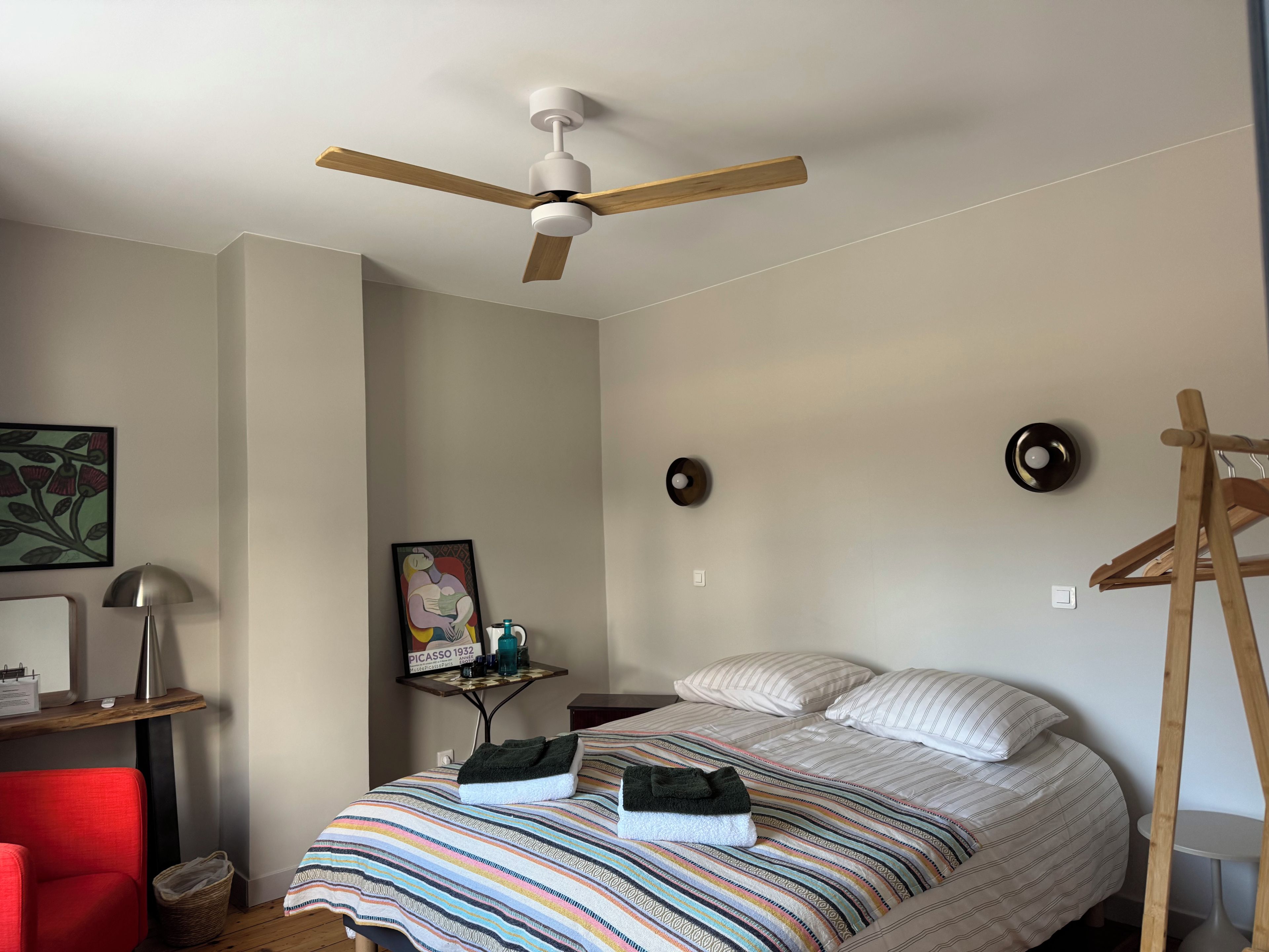 A bedroom with a bed, a ceiling fan, a bedside table with a lamp, a wall-mounted clock, and a framed artwork.