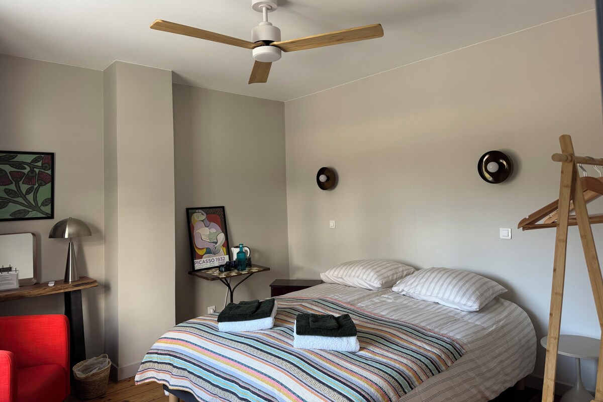 A bedroom with a bed, a ceiling fan, a bedside table with a lamp, a wall-mounted clock, and a framed artwork.