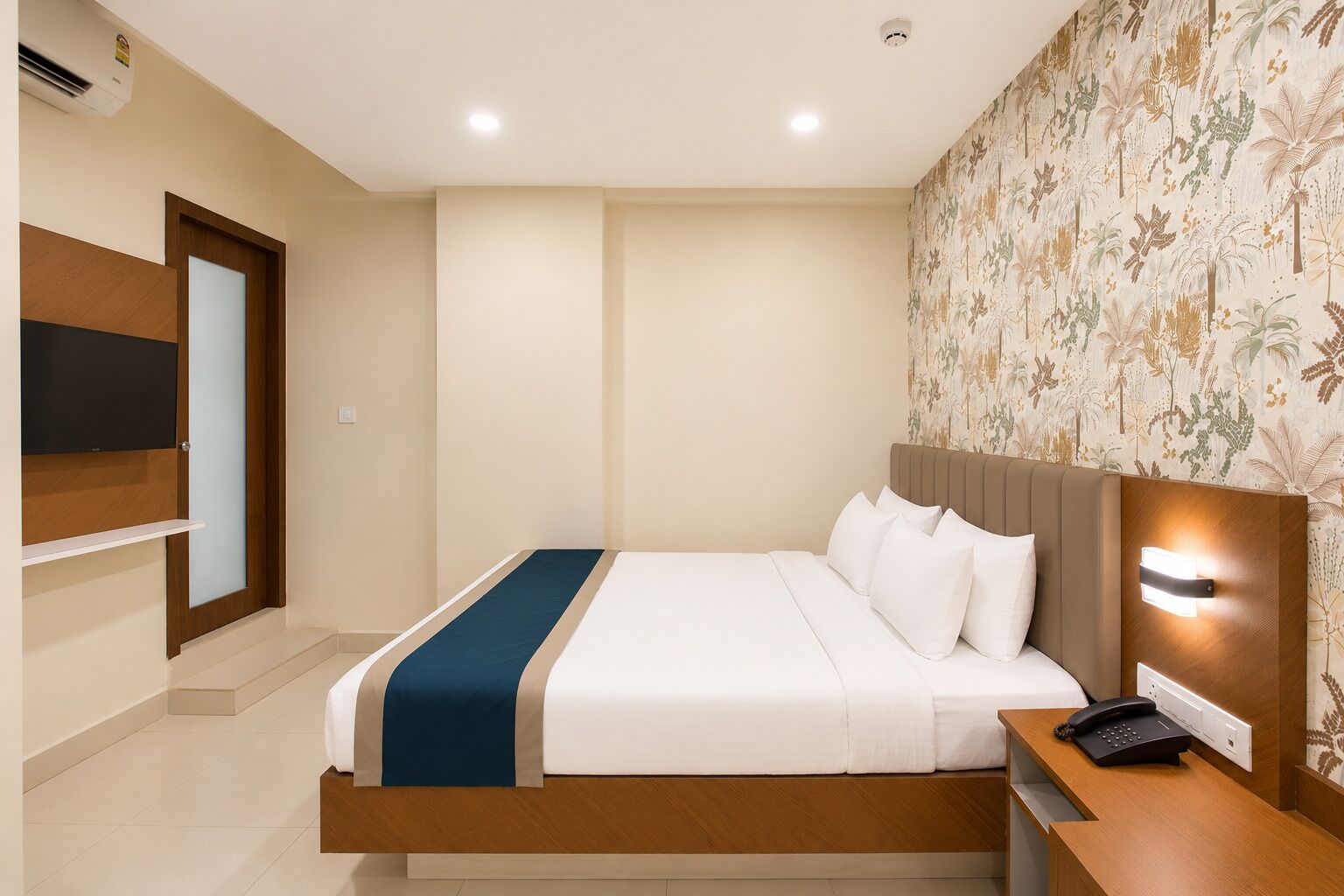 Habitación de hotel con cama, escritorio con teléfono, televisor sobre un mueble y papel tapiz estampado.