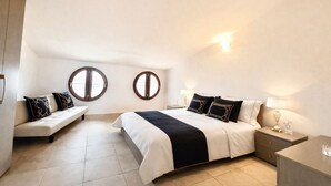 Un dormitorio con una cama, dos mesitas de noche, un banco y dos ventanas circulares fijas en la pared.