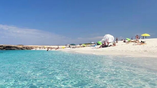 Agua turquesa cristalina, una playa de arena y un edificio blanco a lo lejos.