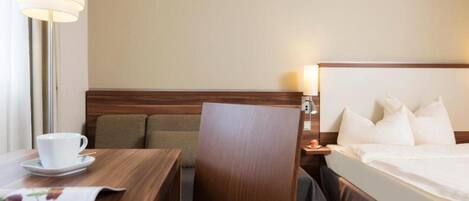 A hotel room with a bed, a desk with a cup and saucer, a chair, and a nightstand with a lamp.