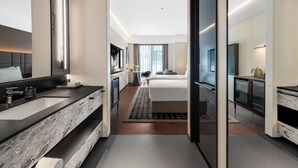 Habitació d'hotel moderna amb un ampli lavabo de marbre, un dormitori independent amb un llit i una taula de nit, i una zona de estar amb un televisor i un sofà.