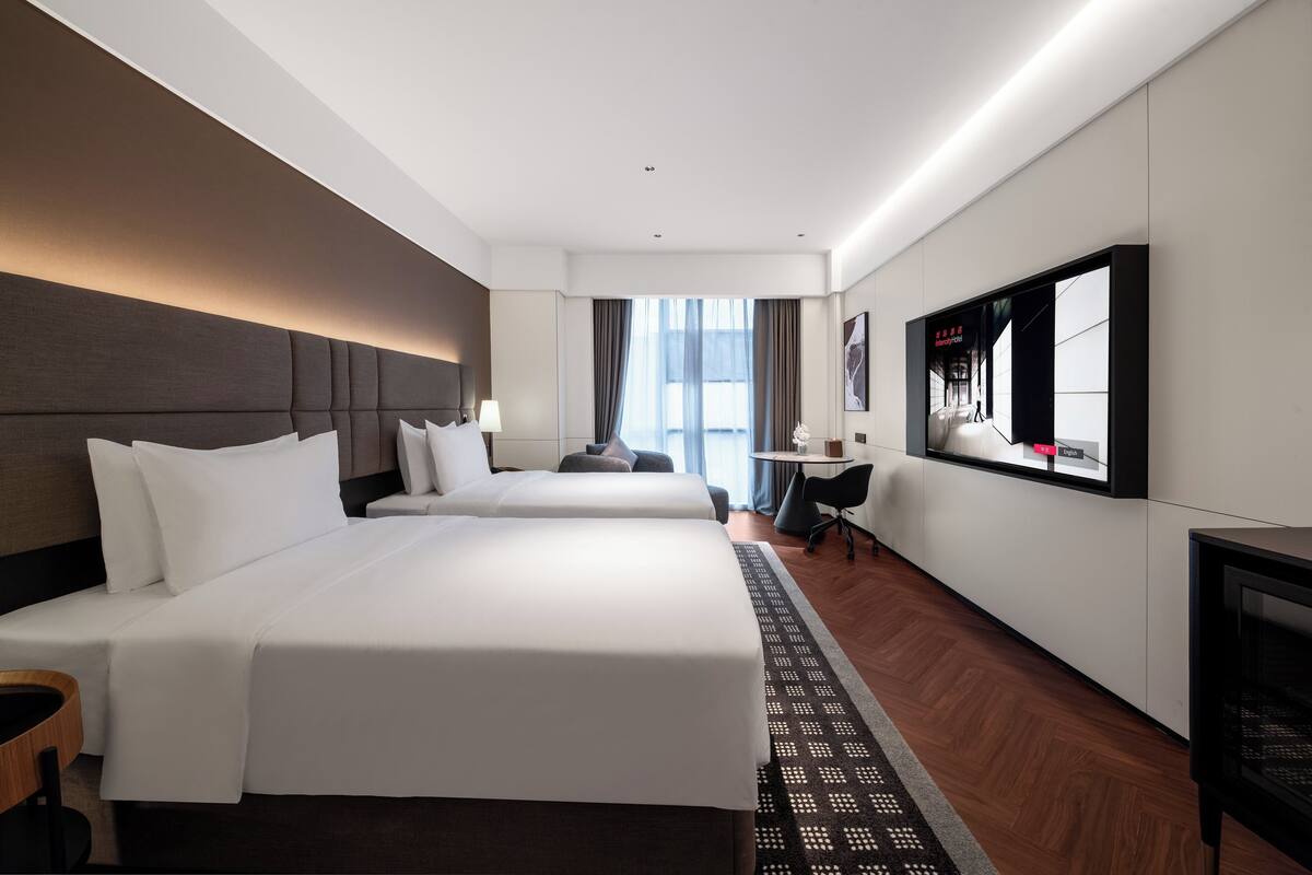 A hotel room with two beds, a desk, a chair, a TV, and a window with curtains.