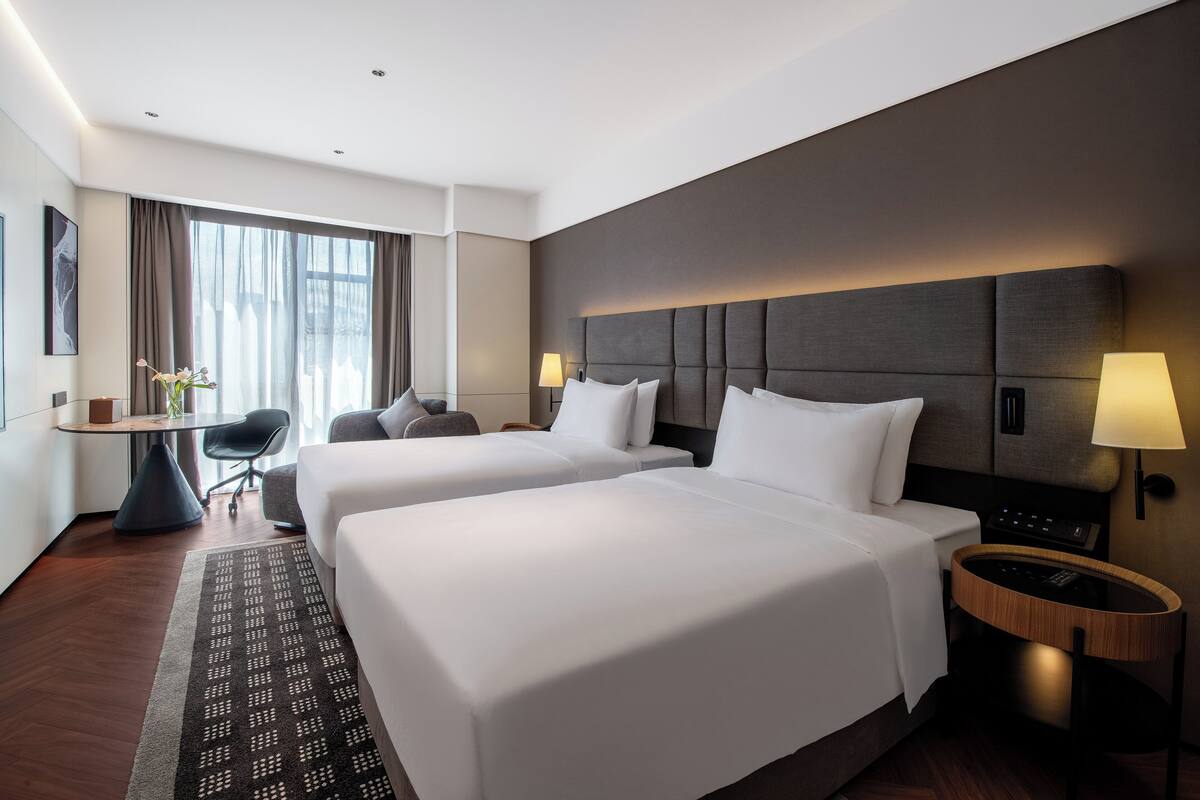 A hotel room with two beds, a desk, a chair, a lamp, and a nightstand.