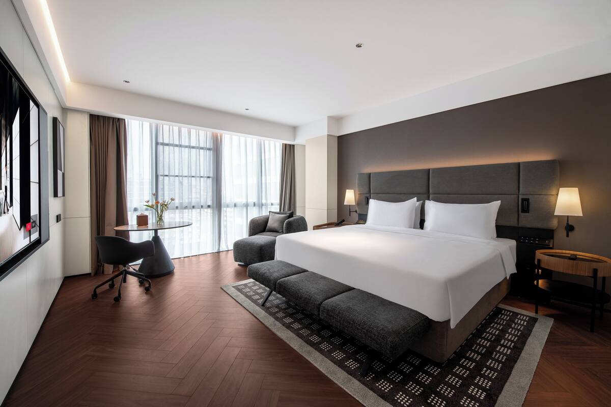 A modern hotel room with a large bed, a desk, a chair, and a view of the city.