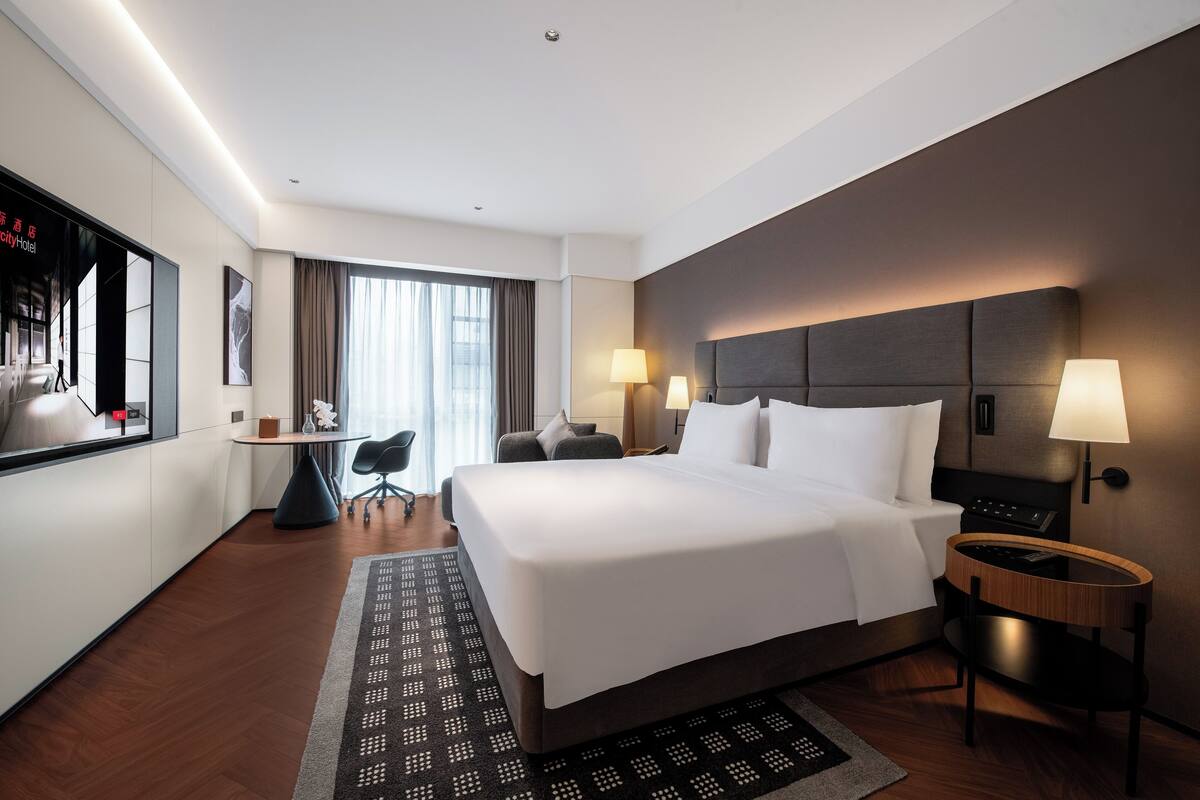 A hotel room with a large bed, bedside lamps, a desk with a chair, and a view of the city.
