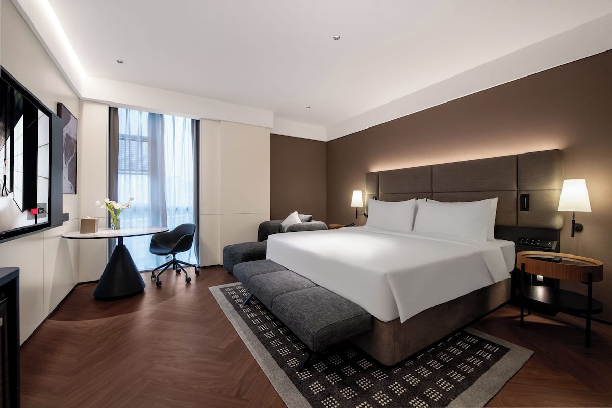 A modern hotel room with a large bed, a desk, a chair, and a view of the city.