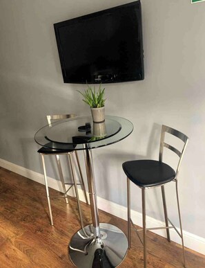 Ein moderner Essbereich mit einem runden Glastisch, zwei Metallstühlen, einer Topfpflanze und einem an der Wand befestigten Fernseher.