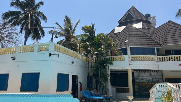 A two-story building with a swimming pool, palm trees, and a person relaxing on a lounge chair. - Luxurious 3-Bedroom Holiday House with Pool Access in Diani Beach (Diani Beach)