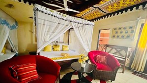 A room with a bed, a red sofa, a wicker chair, a small table with a vase of flowers, and a ceiling fan. - Luxurious 3-Bedroom Holiday House with Pool Access in Diani Beach (Diani Beach)
