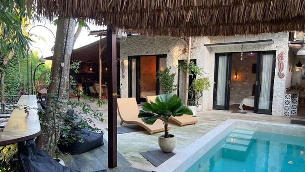 A pool area with lounge chairs, a thatched roof, and a stone building with a bedroom visible. - Cozy Villa El BamboG with a private pool (Blahbatuh)