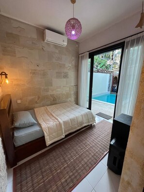 A bedroom with a bed, a wall-mounted air conditioner, a hanging light fixture, and a view of a swimming pool through a glass door. - Cozy Villa El BamboG with a private pool (Blahbatuh)