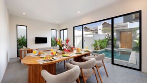 A modern dining area with a wooden table set for breakfast, surrounded by chairs, and overlooking a swimming pool. - Beachside Luxury 3BR Tropical B (Denpasar)