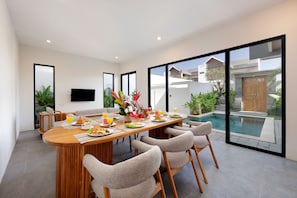 A modern dining area with a wooden table set for breakfast, surrounded by chairs, and overlooking a swimming pool. - Beachside Luxury 3BR Tropical B (Denpasar)