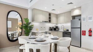 A modern kitchen with white cabinets, a stainless steel refrigerator, and a dining area with white chairs and a round table set for four. - StayShort - Modern 1BR Retreat in MJL Sleeps 4 (Madinat Jumeirah Living)